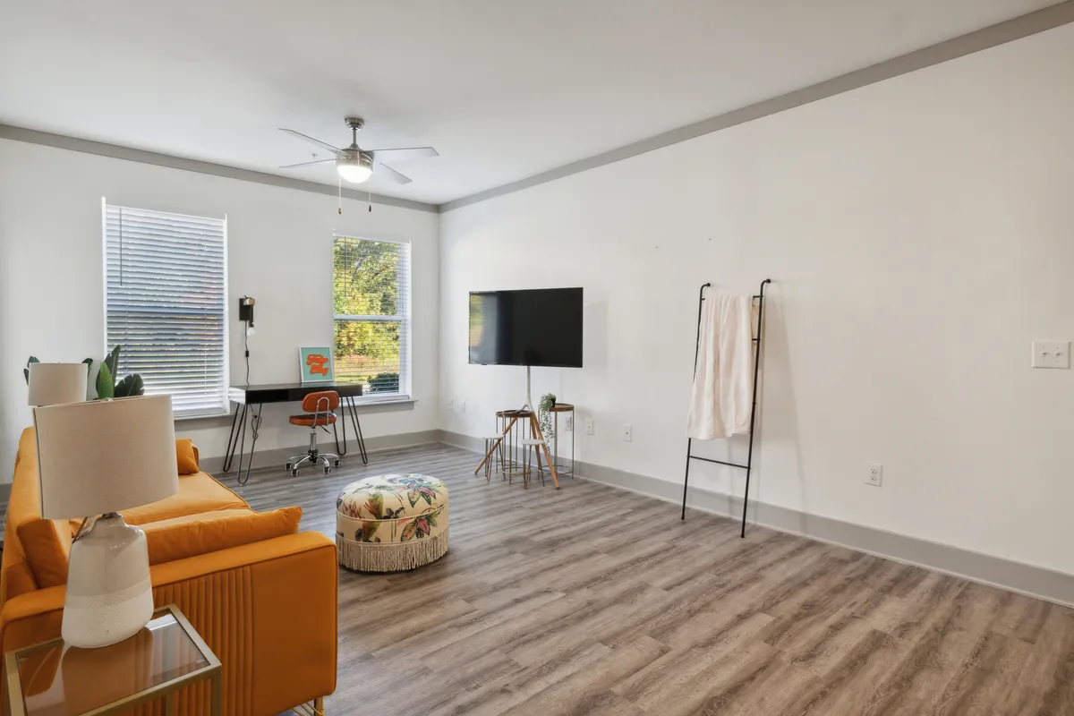 A comfortable and modern living space centered on an orange sofa and colorful pouf, with a stylish desk and chair for working, a TV for entertainment, and a window that lets in abundant natural light. The overall mood is cozy and inviting, ideal for both relaxation and productivity.
