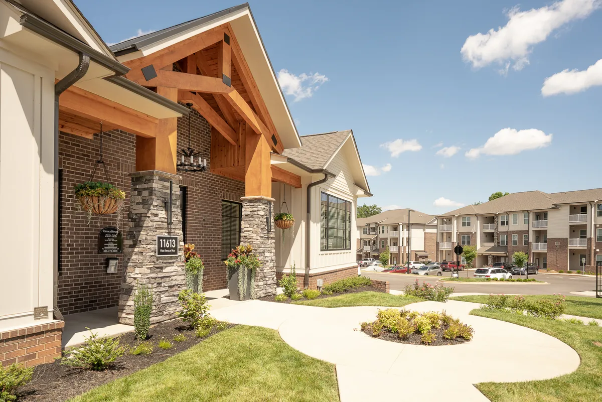 A striking exterior view of a modern apartment building featuring wood beams, stone accents, and lush landscaped gardens. The image captures the inviting entrance and a well-maintained pathway, suggesting easy access to the vibrant city beyond.