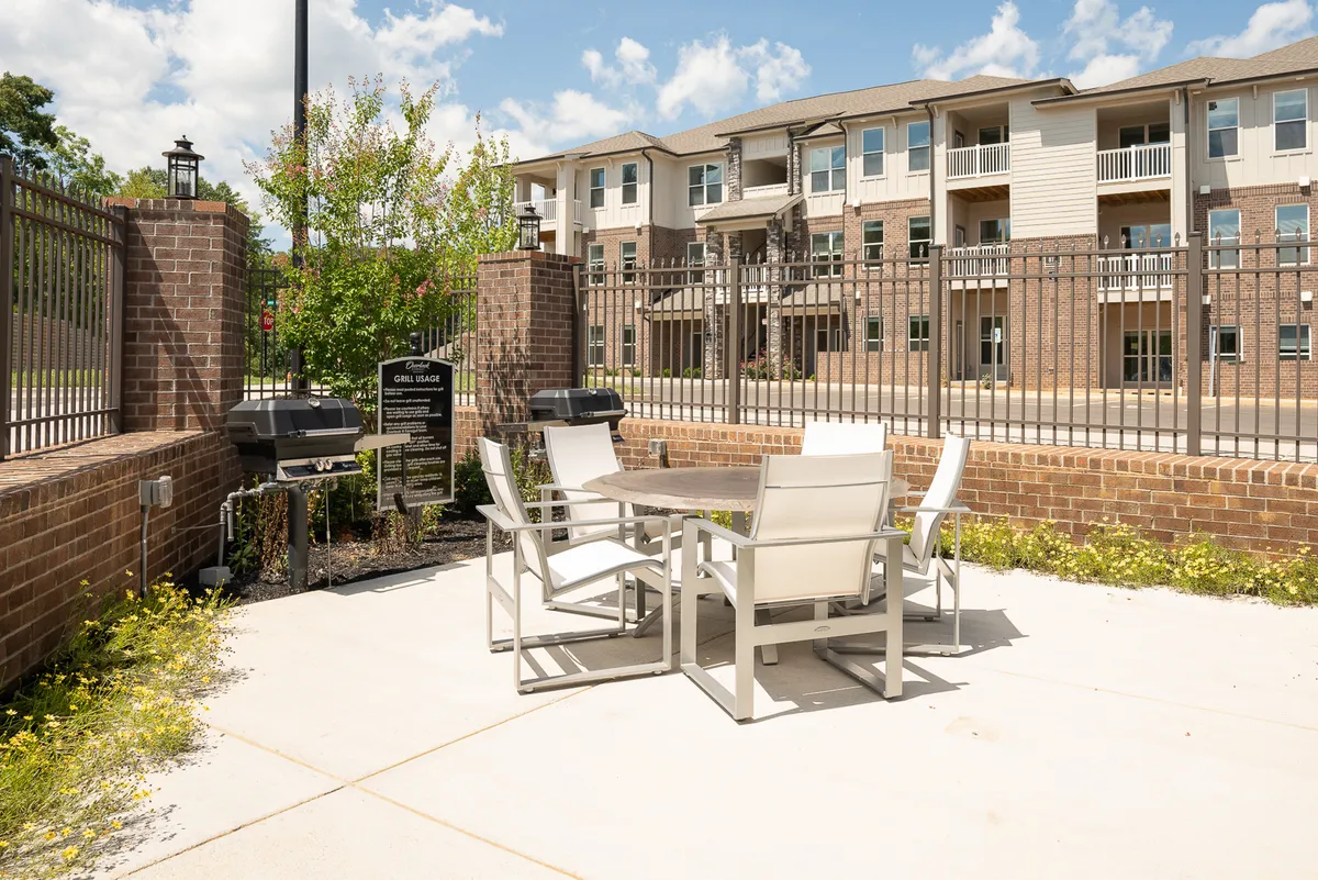 This image features a stylish outdoor dining area, including a dining table, grills, and lush surrounding greenery, all enclosed within a fenced space. The scene suggests a communal atmosphere perfect for outdoor meals, gatherings, or relaxing evenings under the stars.