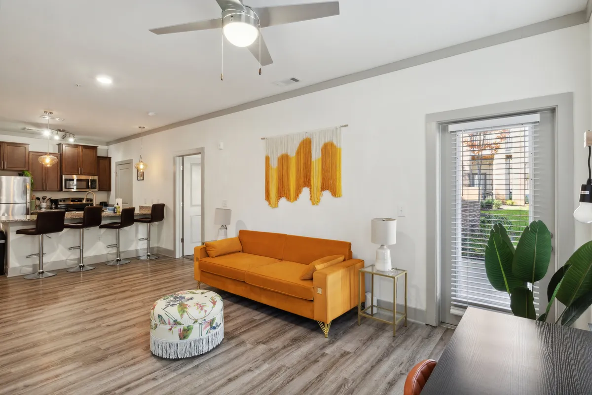 The image showcases a bright, modern living space featuring a vibrant orange sofa as the focal point, complemented by artistic textile wall art above. Sleek bar stools line a kitchen counter, and a small table with a potted plant adds a touch of greenery. The room exudes a cheerful, inviting atmosphere, perfect for relaxation or casual socializing.
