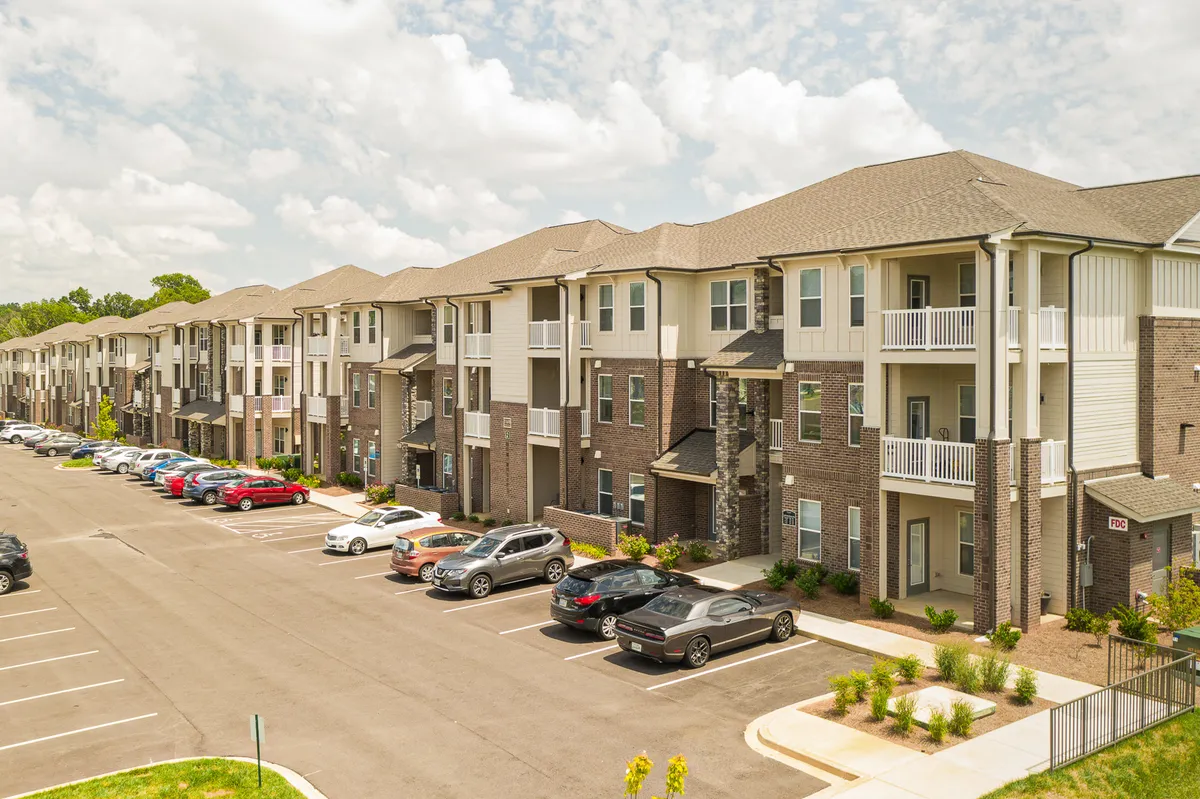 A wide shot of apartment buildings surrounded by green landscaping and parked cars, highlighting communal outdoor spaces where residents can walk and socialize. The image conveys a sense of community and accessibility to city amenities.