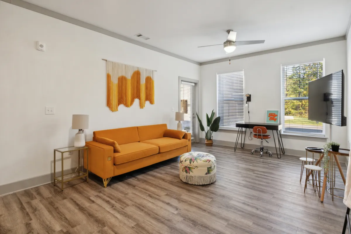 A bright and inviting living room featuring a bold orange designer sofa, a colorful pouf, striking wall art, and a dedicated workspace with a desk and chair. Large windows flood the space with natural light and offer city views, while modern décor and furnishings create a lively yet relaxing environment.