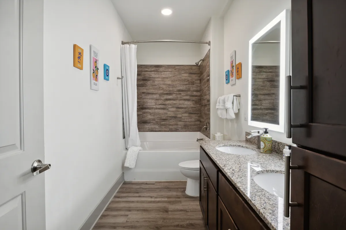 A stylish bathroom featuring double vanity sinks with a granite countertop, a large framed mirror, a bathtub with a shower curtain, and wood-textured wall accents. The space is fresh, inviting, and ideal for both daily routines and relaxation.
