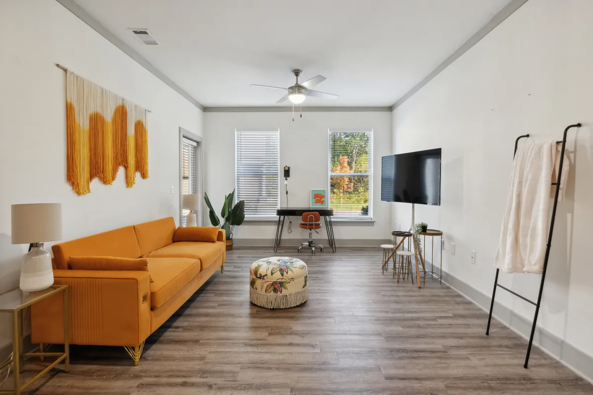 A lively living space with an orange sofa and colorful pouf, stylish wall art, a work desk and chair, and a TV, all set in a room filled with creative energy. The area feels both cheerful and functional, blending spaces for work, leisure, and socializing.