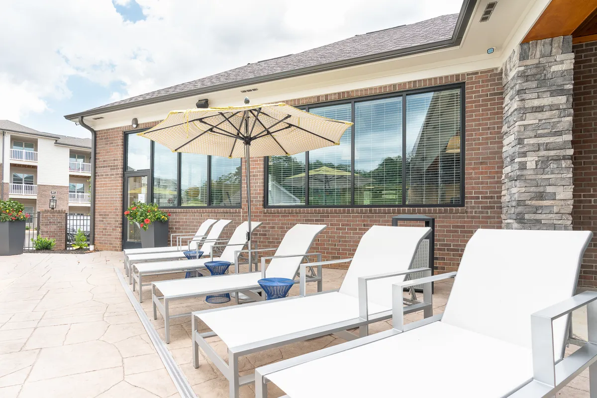 A stylish rooftop area featuring comfortable lounge chairs under umbrellas, decorative flower pots, and a brick wall backdrop. The scene captures people sunbathing, socializing, and enjoying refreshments as the sun sets, emphasizing the inviting atmosphere and panoramic city views.